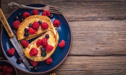 Pancakes with blueberries and raseberries on blue plate on wooden desk.