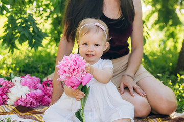 Young pretty mother with daughter lie on a plaid in the park. Family outdoor recreation. Family summer picnic in the park. Mother and daughter outdoors surrounded by pink peonies.
