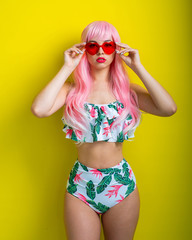 Beautiful girl in a pink wig and colored bikini posing on a yellow background. Woman with artificial long hair and pink glasses in the form of hearts.