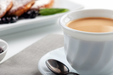 A Cup of coffee with milk and a plate with a pancakes is on the table.