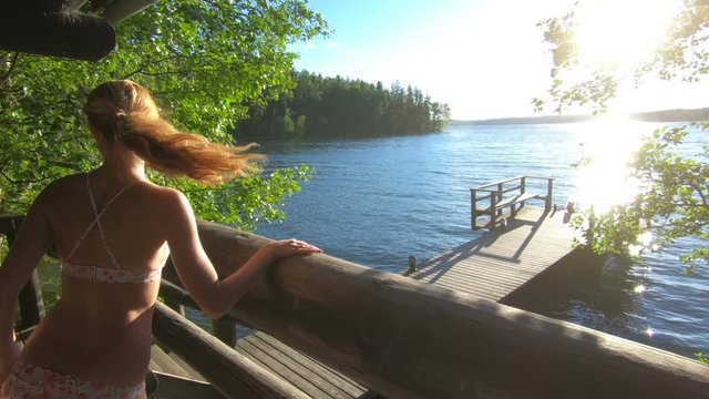 A young slender woman running down the stairs from the sauna to the wooden pier to jump into the lake in summer white night in Finland.