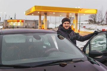 Man holds a cup of coffee standing in front of his car. Patrol station blurred in the background. The concept of coffee pause and rest on the road.