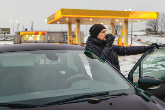 guy drinks a cup of coffee at a gas station in winter standing behind the open car door. The concept of coffee pause and rest on the road.