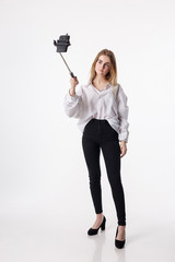 cute girl in shirt, jeans makes self portrait with smartphone attached to selfie stick. young pretty blond female with long hair poses using cell phone isolated on white background. teen communication