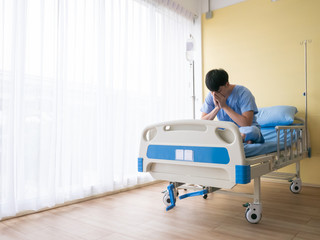 30s Asian patient man handsome over thinking, stressful and suffer about surgery decision alone from accident or diseases sitting on bed while wear blue uniform cloth in VIP room at the hospital