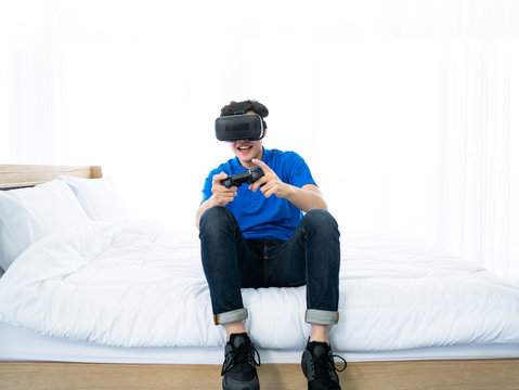 20s Asian young man fun and enjoy from playing virtual reality headset with joystick controller digital technology device for simulation futuristic visual world in glasses with friends in his bedroom