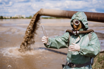 Naklejka premium A male environmentalist in a protective suit and gas mask takes a sample of water. Scientist doing toxicological study.