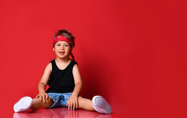 Fototapeta na wymiar Small boy in stylish casual clothing, hair bandana and white sneakers sitting on floor and feeling excited with hands raised up