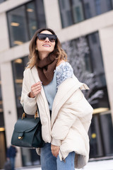 Caucasian model girl in a beige oversized down jacket, knitted sweater with a handbag and glasses...