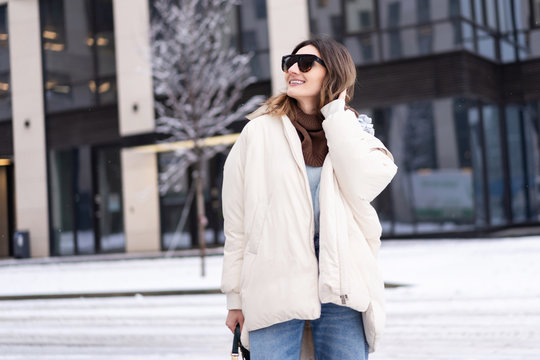 European model in a beige oversized down jacket, knitted sweater with a handbag and glasses posing near the office building. Lifestyle. Female image
