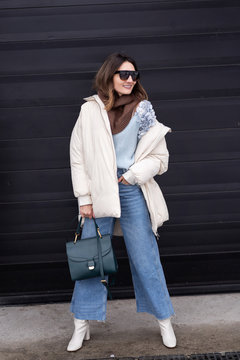 European girl in a beige oversized down jacket, knitted sweater, jeans flared with a handbag and glasses is standing on the street. Life style