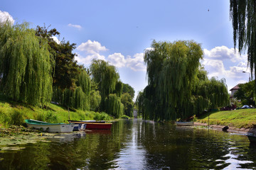Łódki przy brzegach wąskiej rzeczki, zwisające gałęzie wierzby i chmury na błękitnym niebie. © W Korczewski