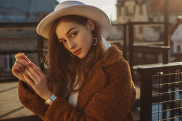 Poster Outdoor fashion portrait of young elegant fashionable brunette woman, model wearing stylish white hat, wrist watch,  brown faux fur coat, posing at sunset, in European city. Copy empty space for text © Victoria Fox