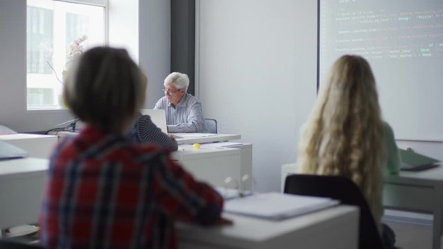 Pan right view of old teacher sitting at table explaining lesson to children pointing at projector screen showing computer code to students learning programming in school class