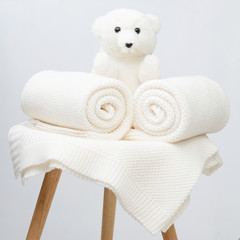 A rolled up knit white plaids. A white teddy bear sits next to it.