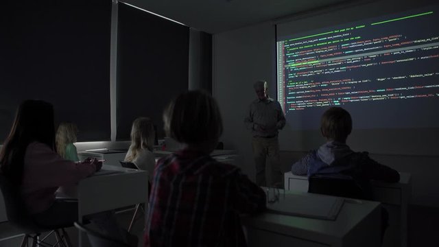 Tilt down view of mature teacher talking to audience while teaching kids programming during computer science lesson in dark classroom in contemporary school