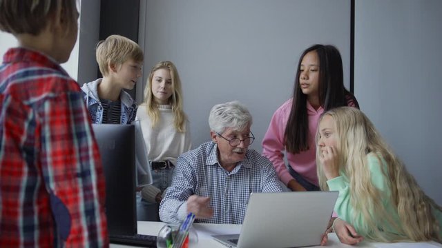 Tilt down of mature male teacher sitting at table with laptop and computer and group of diverse pupils standing around and discussing study project pointing at monitor during computer science lesson