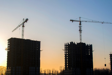 Silhouettes of tower cranes constructing a new residential building at a construction site against sunset background. Renovation program, development, concept of the buildings industry.
