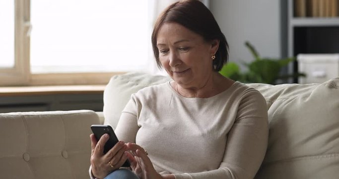 Smiling senior lady using smartphone texting message sit on couch