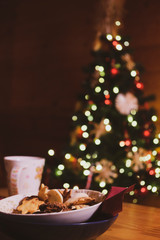 Wooden table set with a bowl of cookies and a mug of hot cocoa, with a decorated Christmas tree and lights, out of focus, in the background.