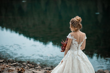 Fototapeta na wymiar Cute pretty blonde bride in luxury wedding dress with bouquet. Wedding on the open air. Bride walking around near the place of the wedding ceremony. Back view