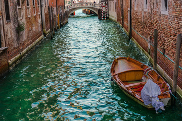 wooden boat floating on the water of a venetian channel between high buildings and a bridge at the end of the channel with some tourists crossing. Horizontal photo