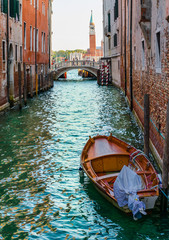 wooden boat floating on the water of a venetian channel between high buildings and a bridge at the end of the channel with some tourists crossing. The tower of Saint Maggiore church at the background