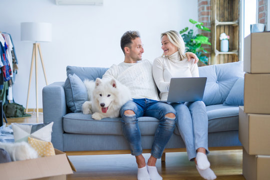 Young beautiful couple with dog sitting on the sofa using laptop at new home around cardboard boxes
