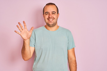 Young man wearing blue casual t-shirt standing over isolated pink background showing and pointing up with fingers number five while smiling confident and happy.