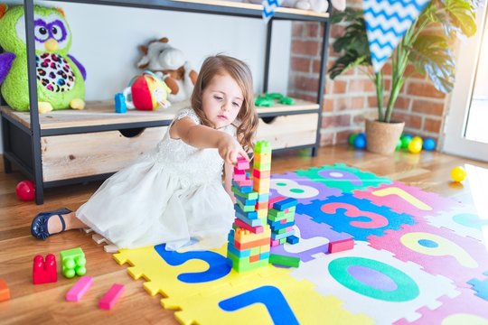 Adorable blonde toddler playing with building blocks toy around lots of toys at kindergarten