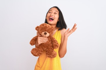 Young beautiful chinese woman holding teddy bear standing over isolated white background very happy and excited, winner expression celebrating victory screaming with big smile and raised hands