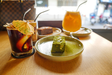 View of one piece of minimal matcha brownie on ceramic dish and cold black coffee with coconut on wooden table and blur background of other chocolate brownie and sunny atmosphere outside cafe. 