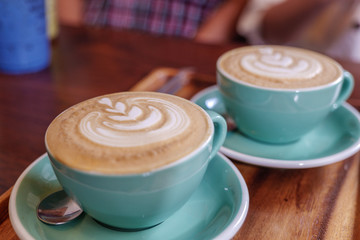 Close up  and selected focus view of two cups of cappuccino with small heart and leave pattern of latte art in pale green ceramic cups on wooden tray and blur background of sitting cozy customer.