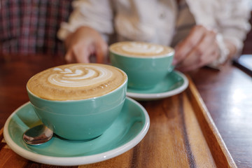 Close up  and selected focus view of two cups of cappuccino with small heart and leave pattern of latte art in pale green ceramic cups on wooden tray and blur background of sitting cozy customer.