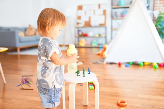 Adorable toddler holding feeding bottle around lots of toys at kindergarten