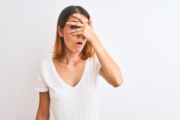 Beautiful redhead woman wearing casual white t-shirt over isolated background peeking in shock covering face and eyes with hand, looking through fingers with embarrassed expression.