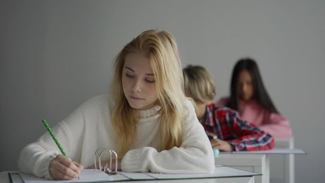 Clever girl with blond hair writing down data while sitting at desk and studying during lesson in school