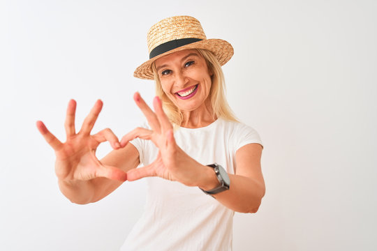 Middle age woman wearing casual t-shirt and hat standing over isolated white background smiling in love showing heart symbol and shape with hands. Romantic concept.