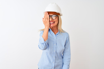 Middle age architect woman wearing glasses and helmet over isolated white background covering one eye with hand, confident smile on face and surprise emotion.