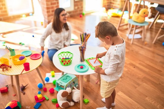 Beautiful teacher and toddler drawing using digital board around lots of toys at kindergarten