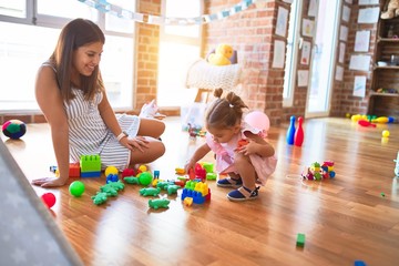 Young beautiful teacher and toddler sitting on the floor playing with building blocks toy at kindergarten