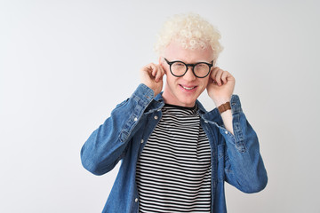 Young albino blond man wearing denim shirt and glasses over isolated white background Smiling pulling ears with fingers, funny gesture. Audition problem