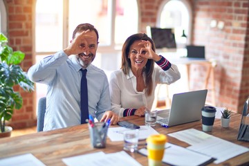 Middle age beautiful business workers working together using laptop at the office doing ok gesture with hand smiling, eye looking through fingers with happy face.