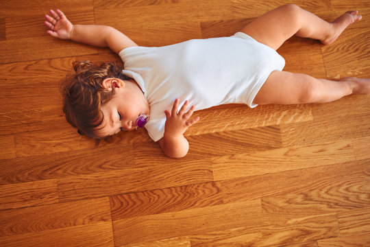 Beautiful toddler child girl wearing white bodysuit lying down on the floor using pacifier