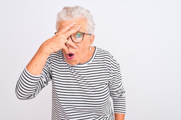 Senior grey-haired woman wearing striped navy t-shirt glasses over isolated white background peeking in shock covering face and eyes with hand, looking through fingers with embarrassed expression.