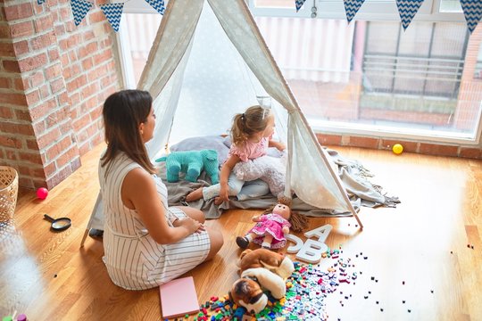 Beautiful teacher and blond toddler girl playing with dolls inside tipi at kindergarten