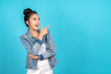 Fototapeta premium Happy asian woman feeling happiness and standing pointing hands to copyspace on blue background. Cute asia girl smiling wearing casual jeans shirt and finger pointing to aside for present promotions.