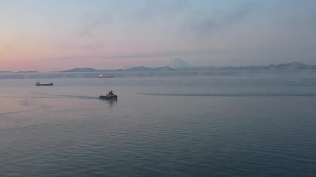 Tug ship at dawn sails across the Pacific ocean on the background of a volcano in Kamchatka, video from a quadrocopter