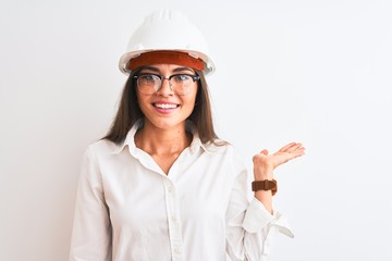 Young beautiful architect woman wearing helmet and glasses over isolated white background smiling cheerful presenting and pointing with palm of hand looking at the camera.