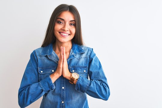Young beautiful woman wearing casual denim shirt standing over isolated white background praying with hands together asking for forgiveness smiling confident.